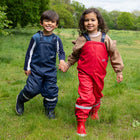 Two children hold hands, smiling in a field, wearing Puddleflex waterproof dungarees: navy for the boy, red for the girl.